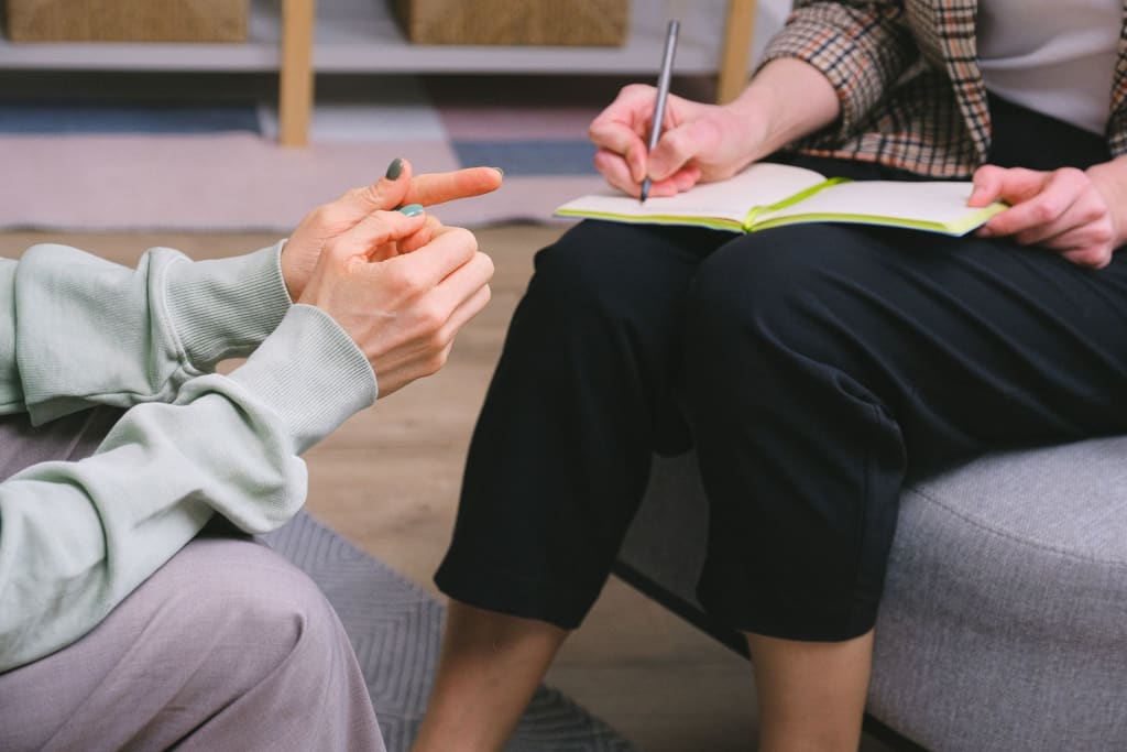 Two people sit opposite each other. One is taking notes in a notebook while the other gestures with their hands.